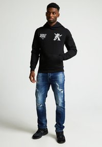 Sudadera negra con estampados gráficos en el pecho, combinada con jeans ajustados de mezclilla claros y desgastados y zapatillas deportivas negras. Fondo simple.