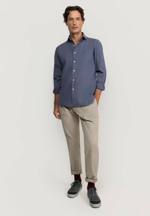 Hombre de pie con las manos en los bolsillos, vistiendo una camisa azul de manga larga, pantalones beige remangados, zapatillas oscuras y calcetines rojos sobre un fondo liso.