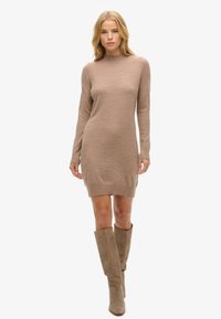 Abito di maglia beige con maniche lunghe e collo alto, caratterizzato da una texture a coste e una silhouette aderente, abbinato a stivali taupe al ginocchio.