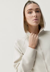 Felpa con zip beige dal design aderente, dotata di un grande cappuccio e dettagli di cucitura visibili. Tessuto liscio e stile contemporaneo.