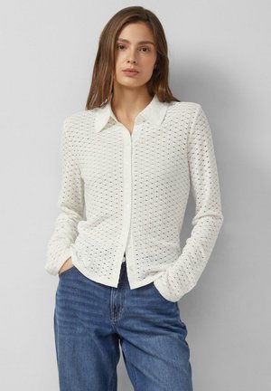 Kobieta w białym swetrze zapinanym na guziki, wykonanym z ażurowej dzianiny, i niebieskich dżinsach, stoi z rękami w kieszeniach na szarym tle.