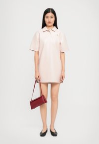 Rochie polo roz deschis cu mâneci scurte, textură netedă și un accent cu logo; asortată cu o geantă structurată de culoare burgundă și pantofi negri cu talpă joasă.
