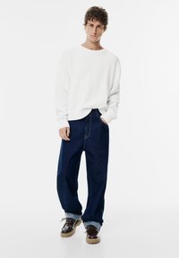 Junger Mann steht mit den Händen in den lockeren, dunkelblauen Jeans-Taschen, trägt einen weißen Rippensweater und braune klobige Schuhe.