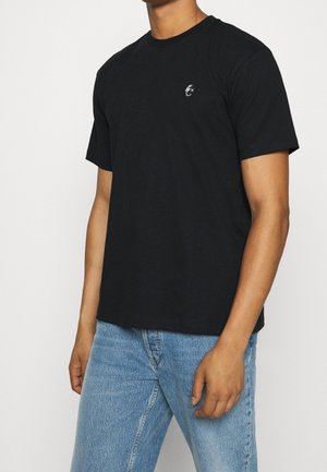 Homme portant un t-shirt noir uni à manches courtes avec un petit logo blanc sur la poitrine, associé à un jean bleu clair, sur fond blanc.