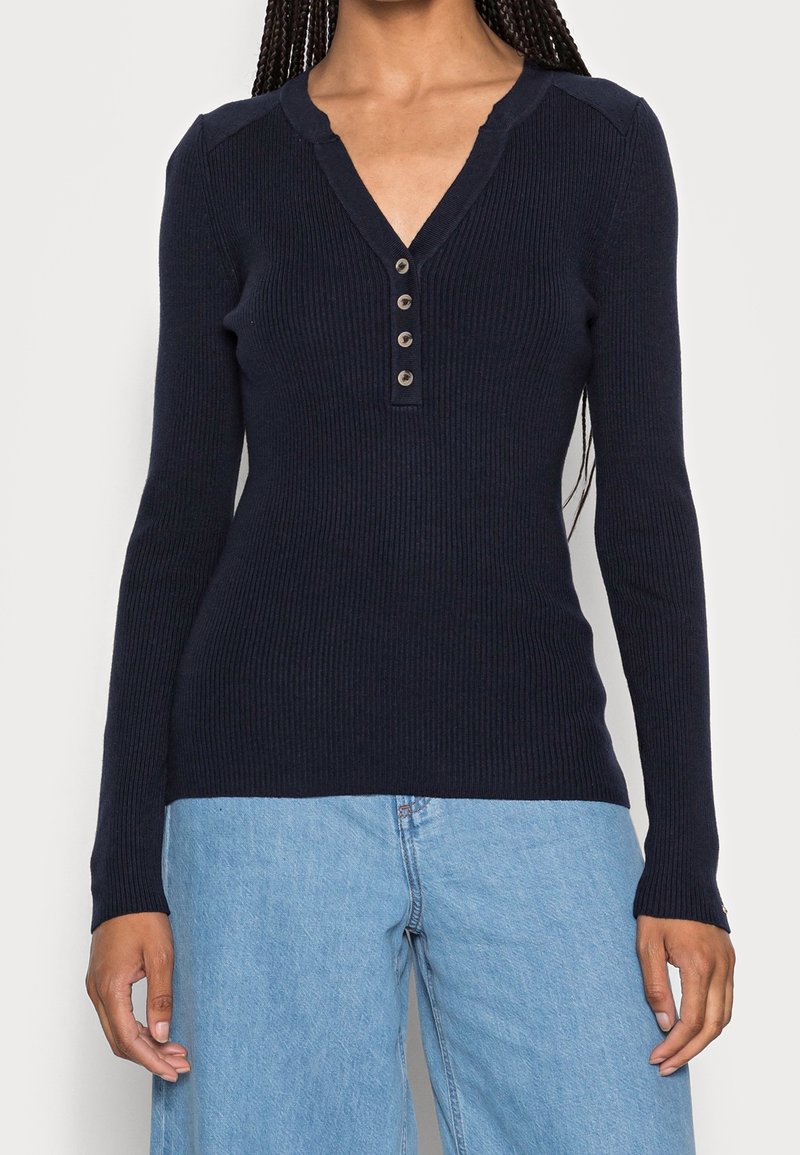 Haut à manches longues côtelé marine avec encolure en V et patte de boutonnage à cinq boutons. Conception ajustée et texture douce, associé à un jean large bleu clair.