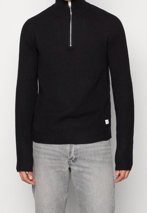 Homme portant un pull en maille côtelée noire avec un col demi-fermeture éclair argentée et un jean gris clair, debout devant un fond uni.