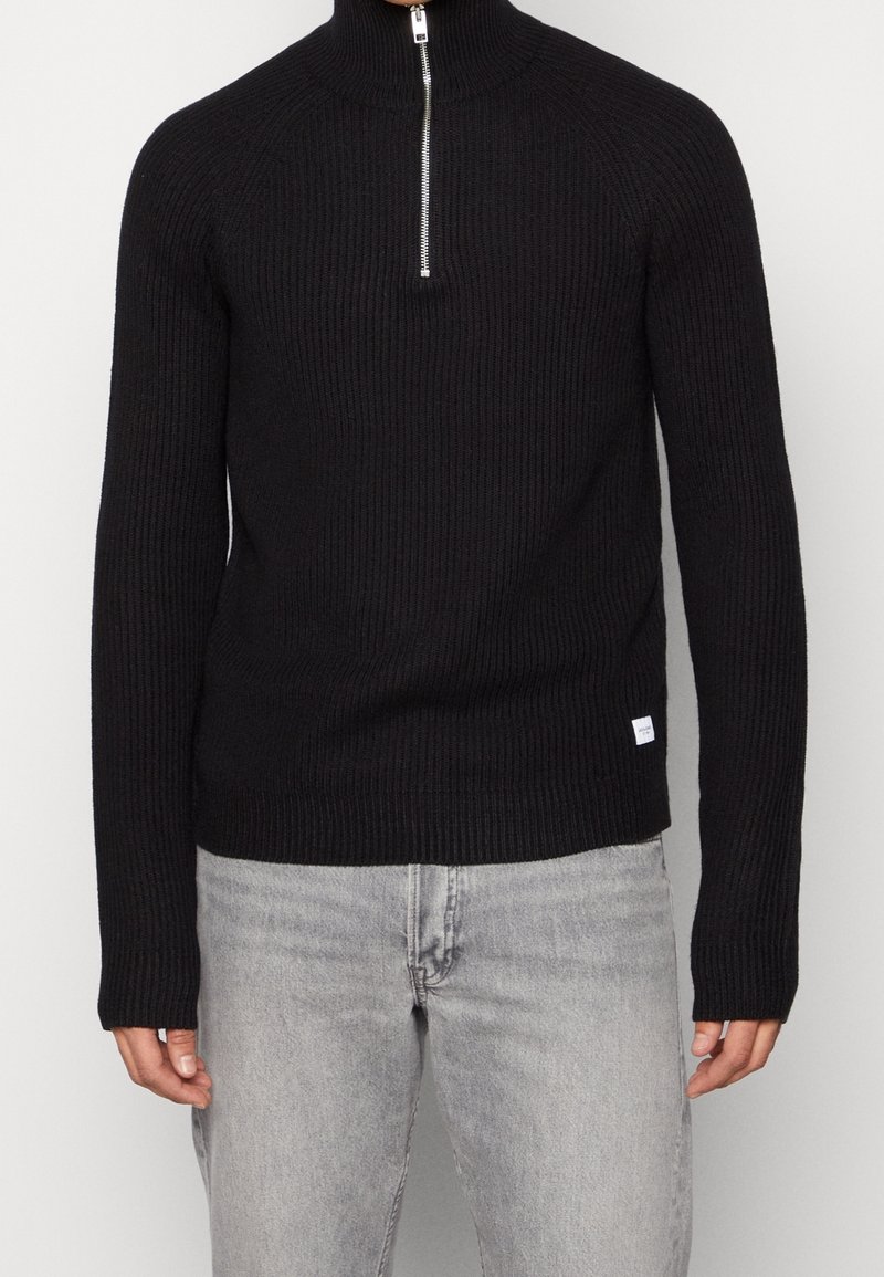 Homme portant un pull en maille côtelée noire avec un col demi-fermeture éclair argentée et un jean gris clair, debout devant un fond uni.