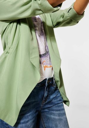 Světlé zelené volné sáčko s ohrnutými rukávy, nošené přes tričko s potiskem a modré džíny. Hladká látka s uvolněným střihem.
