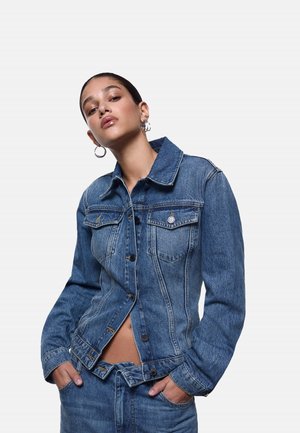 Giovane donna che indossa una giacca di jeans blu e jeans, con le mani nelle tasche, in piedi davanti a uno sfondo chiaro e semplice.