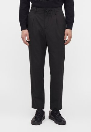 Man die zwarte op maat gemaakte broek draagt met een aantrekkoord in de taille, gecombineerd met zwarte sneakers en een donker shirt met lange mouwen.
