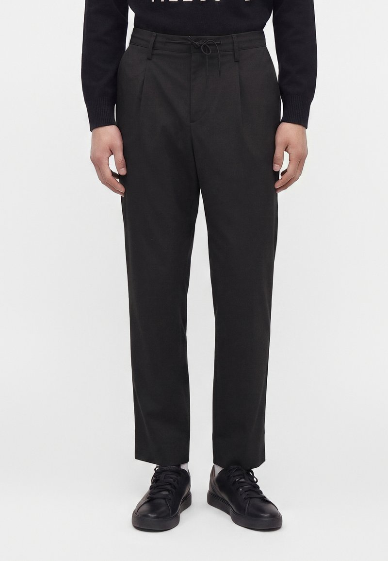 Homme portant un pantalon noir sur mesure avec taille à cordon, associé à des baskets noires et un haut sombre à manches longues.