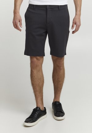 Pantalones cortos negros de tela suave, con cierre de botón, trabillas para cinturón y piernas rectas. Combinados con zapatillas negras.