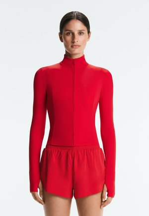 Vrouw die voor zich uit kijkt, draagt een aansluitend rood topo met lange mouwen en rits en rode sportshorts tegen een effen lichte achtergrond.