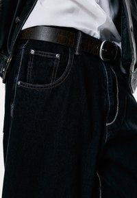 Person iført sorte jeans med detaljeret syning, et brunt tekstureret bælte, en hvid skjorte stoppet i, og en sort læderjakke.