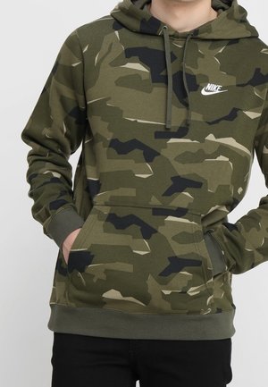 Osoba nosząca zielono-czarną maskującą bluzę z kapturem Nike z rękami w przedniej kieszeni, w połączeniu z czarnymi spodniami.