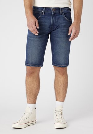 Homme portant un short en denim bleu, des baskets blanches et des chaussettes blanches montant jusqu'à la cheville, se tenant devant un fond uni.