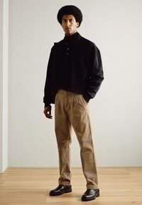 Camisa negra de manga larga con cuello tipo polo, pantalones beiges y botas negras. Tejido suave con un corte holgado. Fondo de suelo de madera.
