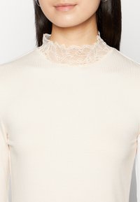 Ribbede langermede topp i lys beige med dekorativt blondehals. Glatt tekstur med en tettsittende silhuett.