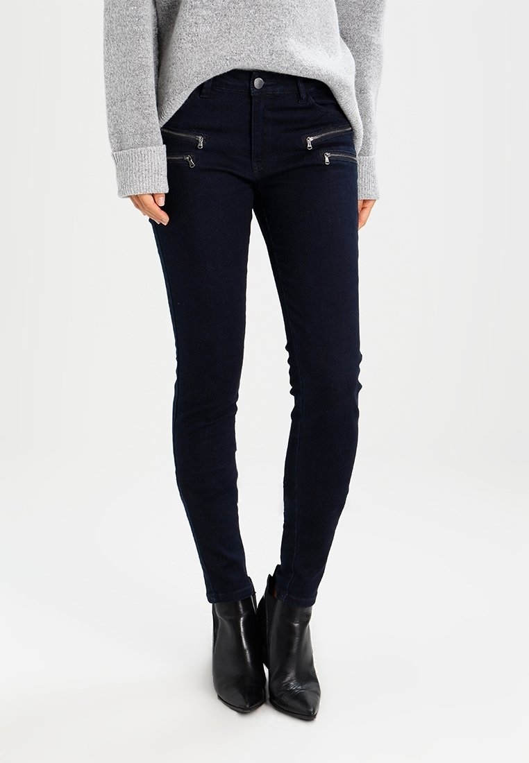 Jeans ajustados de color azul oscuro con bolsillos frontales con cremallera, confeccionados en denim elástico, combinados con botas negras hasta el tobillo y un suéter gris.