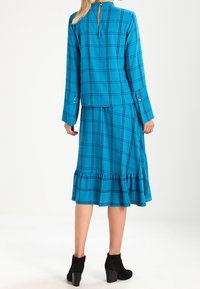 Abito a scacchi teal con gonna svasata, maniche lunghe e dettagli a bottoni. Presenta uno scollo a chiave sul retro e una texture morbida. Stivaletti neri alla caviglia.