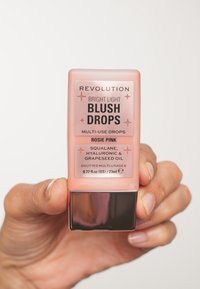 Lysende Blush Dråper i Rosie Pink, oppbevart i en klar glassflaske med en metallisk sølvbase. Inneholder squalan, hyaluronsyre og druefrøolje.