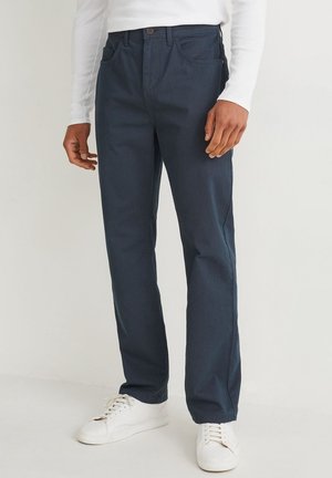 Hombre con pantalones azul oscuro de pierna recta, camiseta blanca de manga larga y zapatillas blancas, de pie contra un fondo liso.