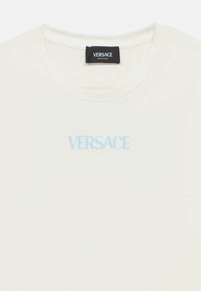 Bavlněné tričko krémové barvy s kulatým výstřihem. Světle modré logo „VERSACE“ vytištěné uprostřed na hrudi. Hladká textura.