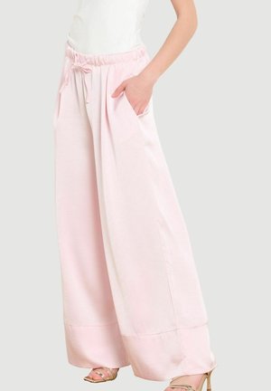 Naine, kes kannab heledat roosa laia säärega pükse elastse vöökoha ja nööriga, käsi taskus, koos kuldsete rihmik-kontsakingadega.