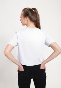 Mladá žena s hnědými vlasy svázanými do culíku, oblečená v bílém tričku a černých kalhotách, rukama v zadních kapsách, odvrácená od kamery.