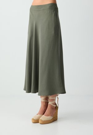 Femme portant une jupe midi trapèze vert olive associée à des espadrilles compensées beiges nouées autour des chevilles.