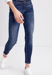 Jeans ajustados de color azul oscuro con cintura alta, ligeros desgastes y detalles de costura dorada, combinados con zapatillas blancas.