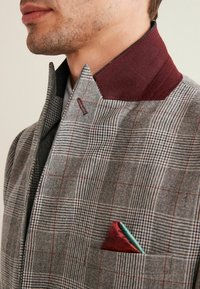 Giacca a quadri grigia con colletto bordeaux, dotata di un fazzoletto da taschino in tessuto rosso e verde. Materiale in lana strutturato, design su misura.
