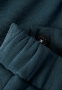 Detaljbild av en mörk teal ribbad tygmanschett med en svart knapp och elastisk detalj inuti för justerbar passform.