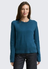 Tuerkiser Wollpullover mit rundem Ausschnitt, gerippten Bündchen und Saum. Kombiniert mit dunklen Jeans, verfügt er über eine glatte Textur.
