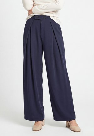 Donna che indossa pantaloni blu navy a gamba larga con pieghe frontali e scarpe beige con tacchi alti, ritratta dal torso, in piedi su sfondo bianco.