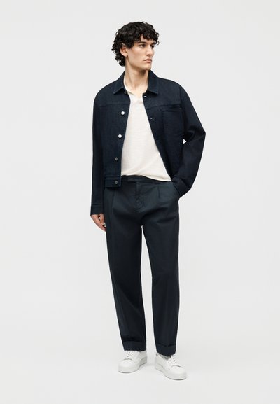 Jeune homme aux cheveux bouclés et foncés portant une veste en denim noire, une chemise blanche, un pantalon noir et des baskets blanches, debout avec une main dans la poche.