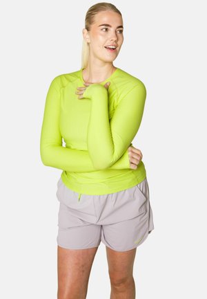 Žena oblečená v jasne zelenej tričku s dlhým rukávom a svetlosivých šortkách, stojaca so založenými rukami a usmievajúca sa, pozerajúc sa do strany.