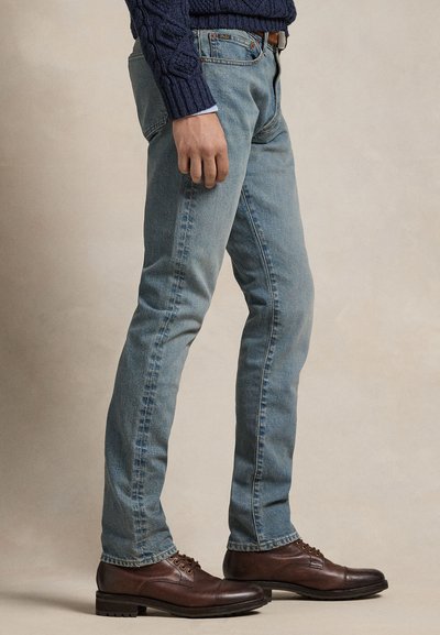 Osoba nosící světle modré džíny slim fit, tmavě hnědé kožené společenské boty a tmavě modrý svetr s copánkovým vzorem, stojící bokem proti jednobarevnému pozadí.