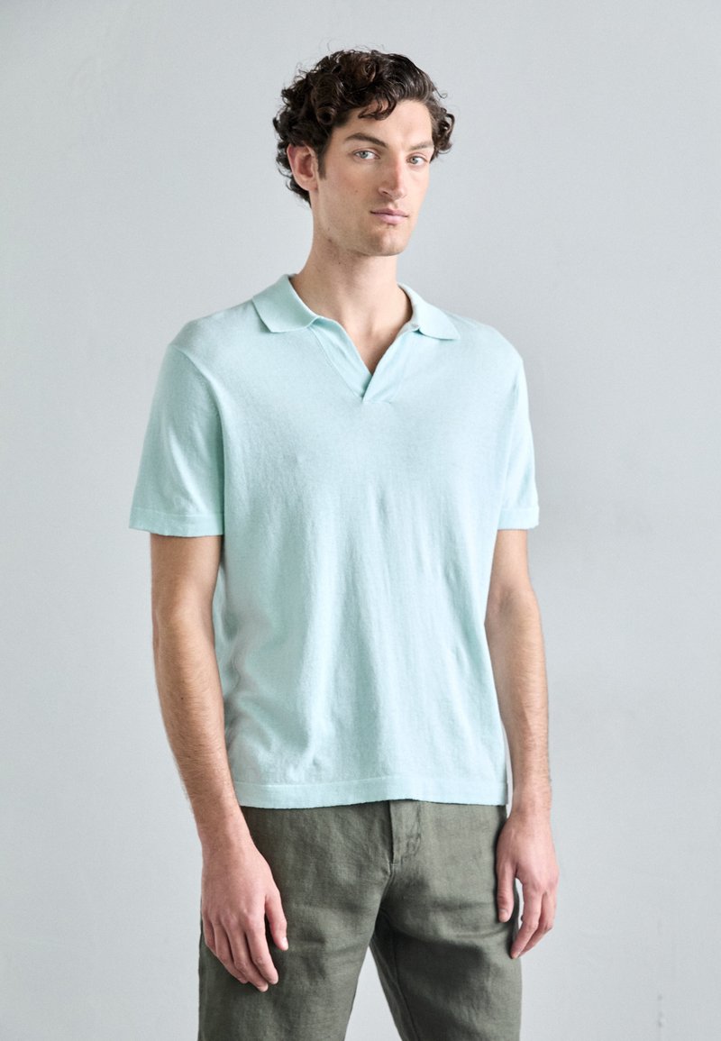 Cămașă polo din bumbac de culoare bleu deschis, cu mânecă scurtă și textură moale, combinată cu pantaloni de culoare verde măsliniu.