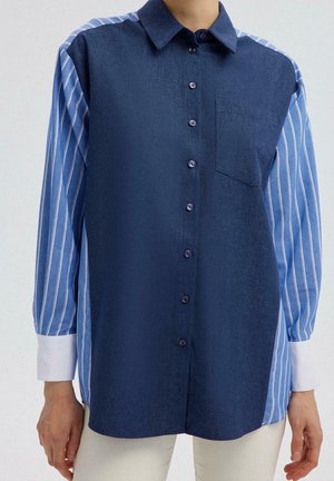 Femme portant une chemise boutonnée avec un devant uni bleu foncé et des manches rayées bleu clair et blanc avec des poignets blancs.