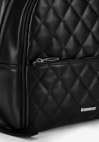 Sac à dos en cuir matelassé noir avec une poche zippée à l'avant, des éléments en métal argenté et une petite plaque logo. Texture lisse avec un motif en diamants.