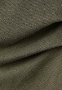 Gros plan d’un tissu texturé vert olive avec un motif fin et serré ainsi que des plis doux créant des ombres douces.