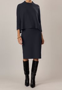 Maglione navy a maglia con maniche a tre quarti, indossato sopra una gonna aderente abbinata. Stivali neri al ginocchio a punta completano l'outfit.