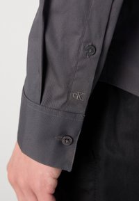 Graues Baumwollhemd mit Knopfmanschette und gesticktem Logo. Der Stoff wirkt glatt mit dezentem Glanz und strukturierter Gestaltung.