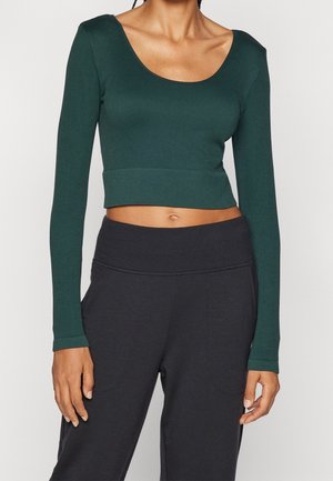 Vrouw draagt een donkergroen cropped top met lange mouwen en een zwarte broek met hoge taille en zichtbare zakken aan de zijkanten.