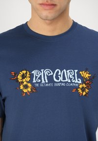 Granatowa bawełniana koszulka z kolorowym wzorem kwiatowym wokół logo "Rip Curl" w białym kolorze, z pomarańczowymi i żółtymi akcentami.