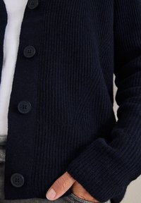Navy gestrickte Strickjacke mit geripptem Muster, großen schwarzen Knöpfen und langen Ärmeln. Das Design umfasst eine Tasche an der unteren Seite.