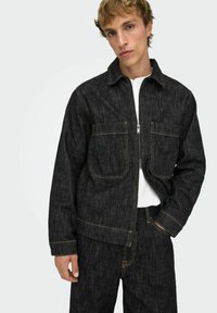 Mustast teksariidest jakk, mille ees on lukk, kaks rinnataskut ja nähtavad kollased õmblused. Kangas tundub tugev ning tekstuuriga viimistlusega.