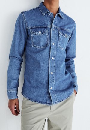 Homme portant une chemise en denim bleu avec boutons et poches poitrine, ainsi que un pantalon beige, debout avec une main dans la poche.
