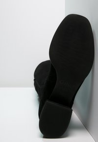 Botín negro de ante hasta el tobillo con suela de goma texturada, con cremallera lateral y detalle de logo en el tacón. Acabado suave, sin patrones visibles.